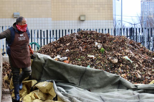 A worker wearing an apron and a bandana over his face stands near a large pile of compost at the Big Reuse site under the Queensboro Bridge.